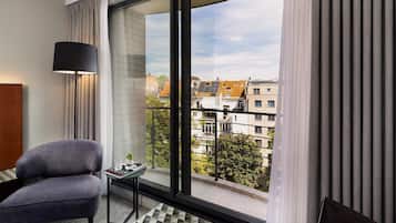 A modern hotel room with a large window, a grey armchair, a small table, and a floor lamp.