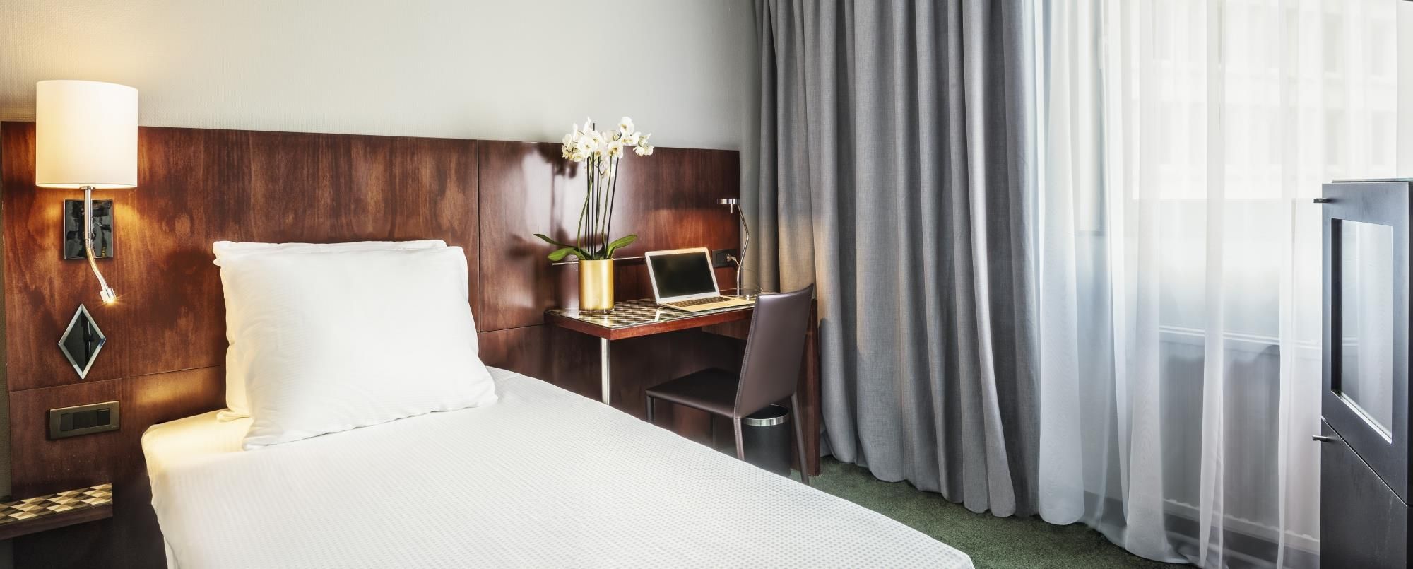 Habitación de hotel con cama, escritorio, computadora portátil, silla, lámpara y ventana con cortinas.
