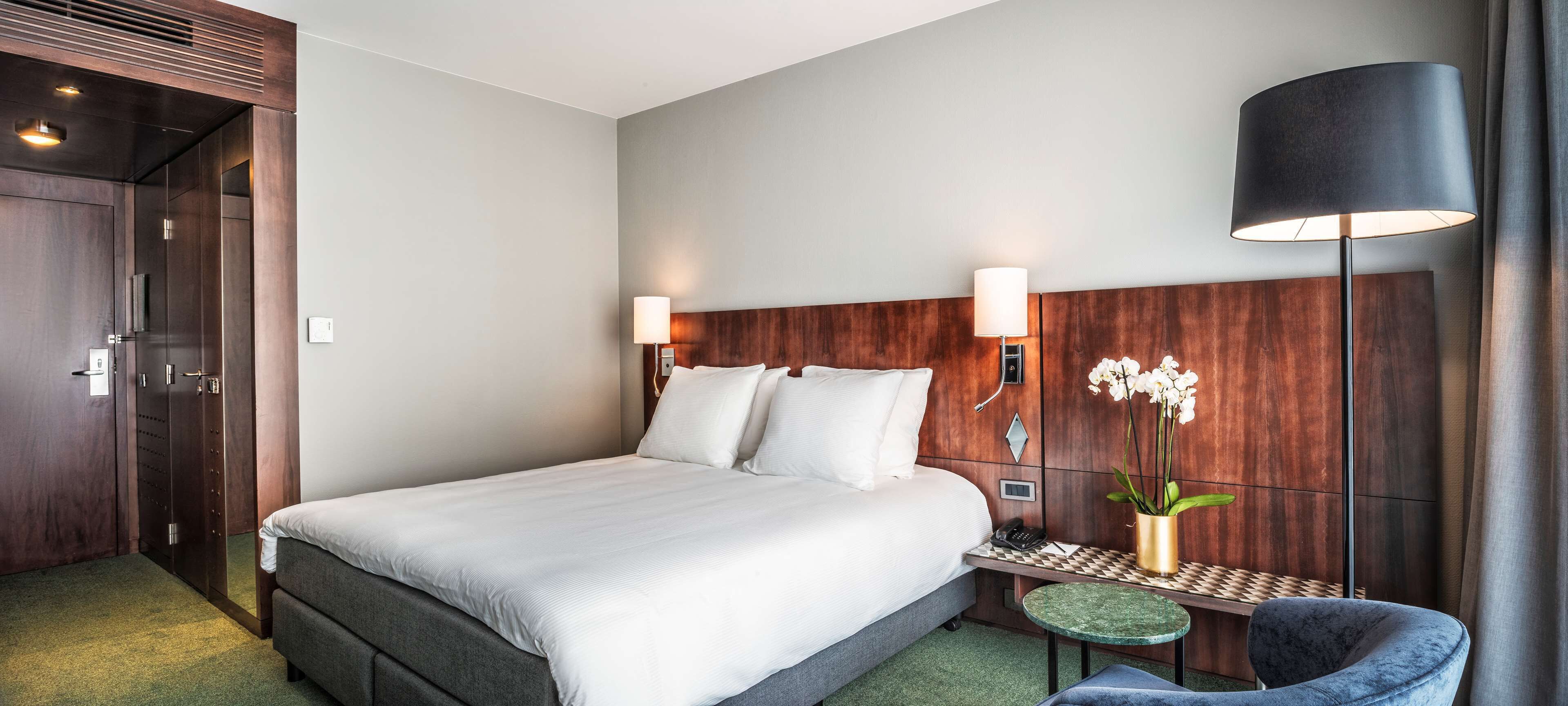 A hotel room with a large bed, a bedside table with a lamp, a small round table, a chair, and a wooden headboard.