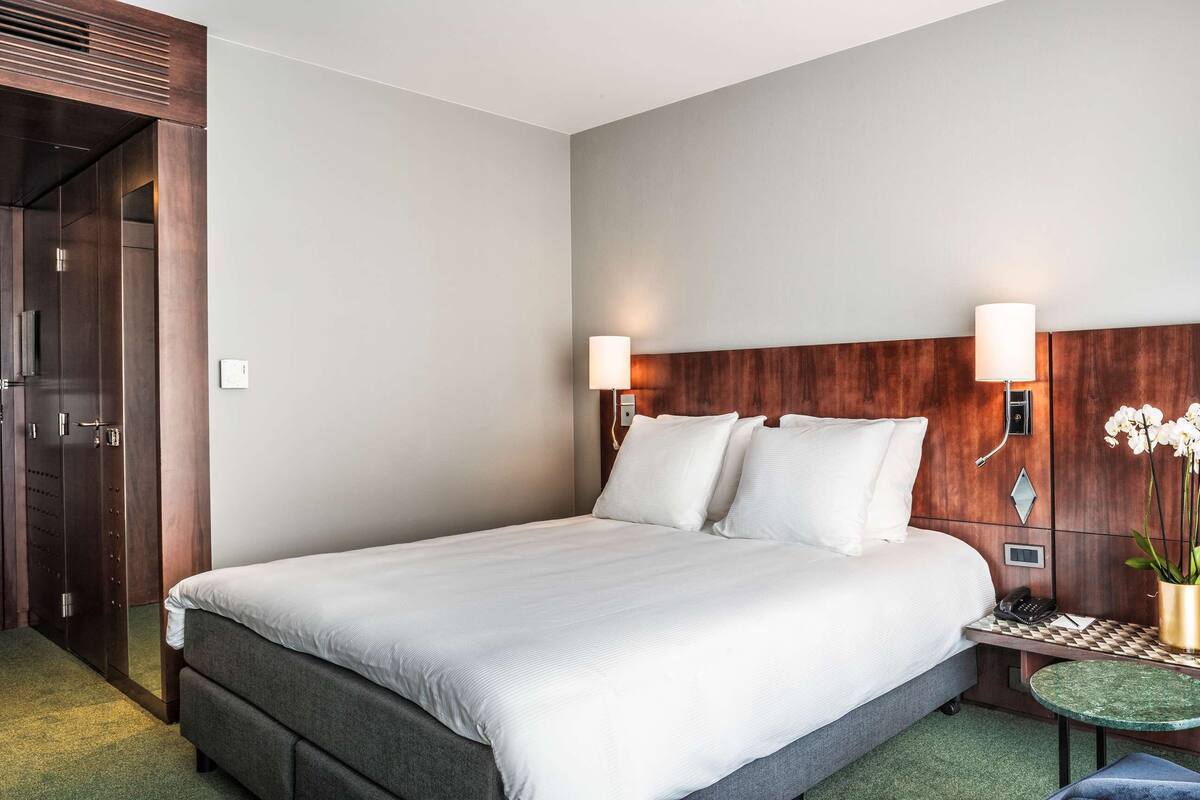 A hotel room with a large bed, a bedside table with a lamp, a small round table, a chair, and a wooden headboard.