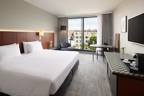 Habitación de hotel con una cama grande, un escritorio con cafetera, una silla y vistas a edificios por la ventana.