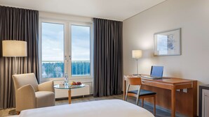 A hotel room with a bed, a desk with a laptop, a chair, a table with fruit, and a window with curtains. - Dorint Am Nürburgring Hocheifel (Nuerburg)