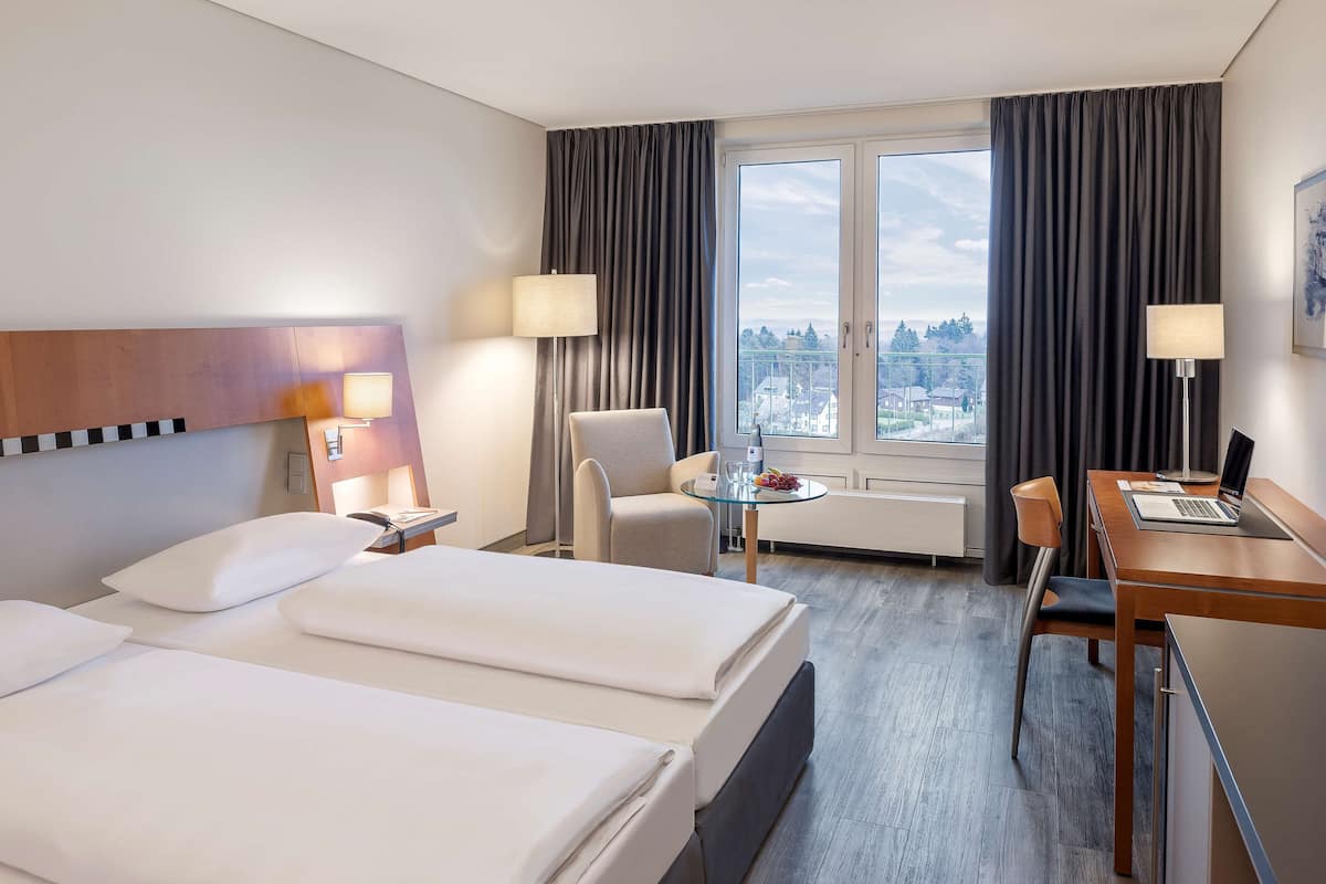 Habitación de hotel con dos camas, un escritorio, una silla, una mesita y vistas a la ciudad a través de amplias ventanas.