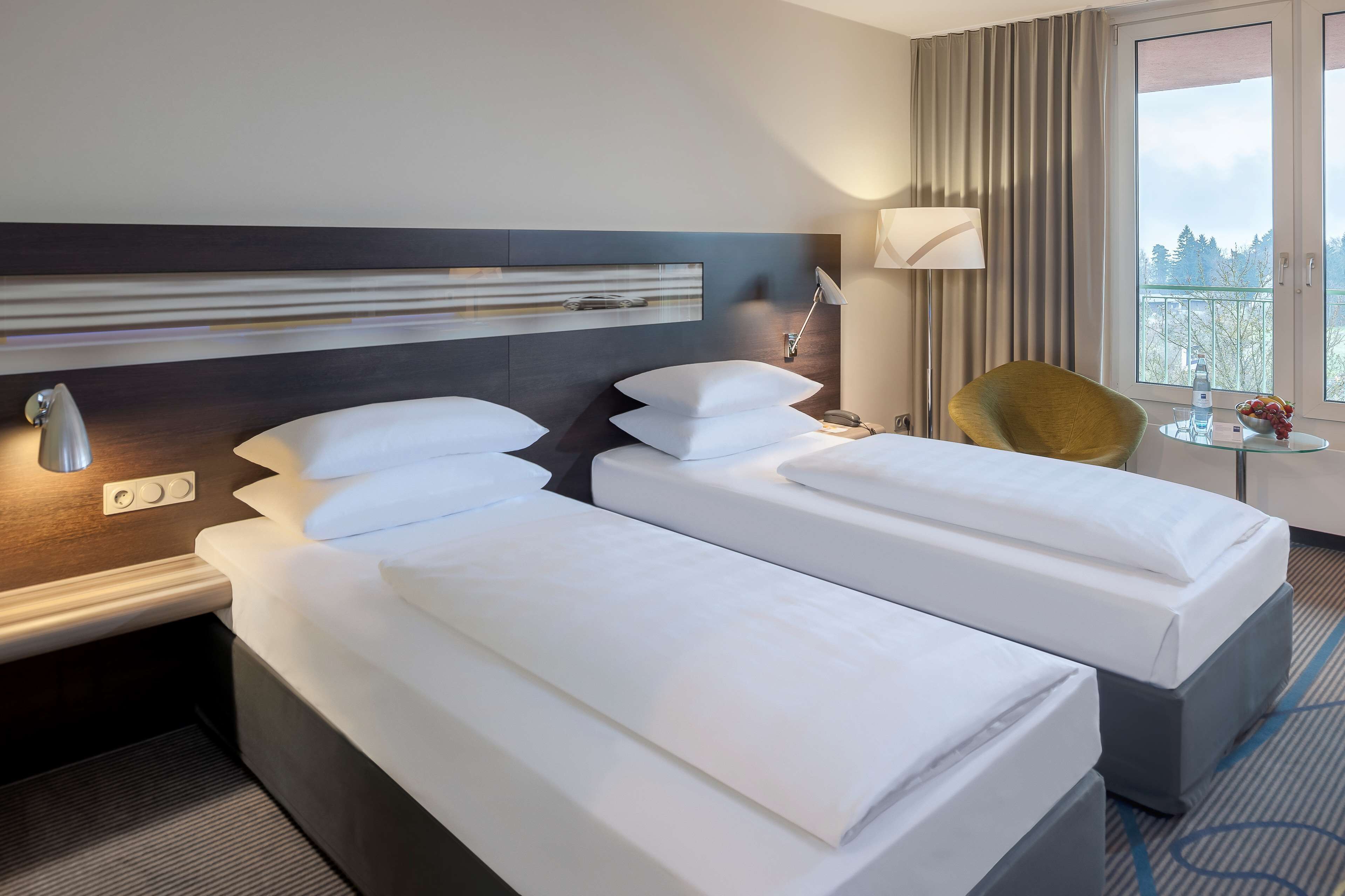 A hotel room with two beds, a modern headboard, a small table with a lamp, and a view of the outdoors.