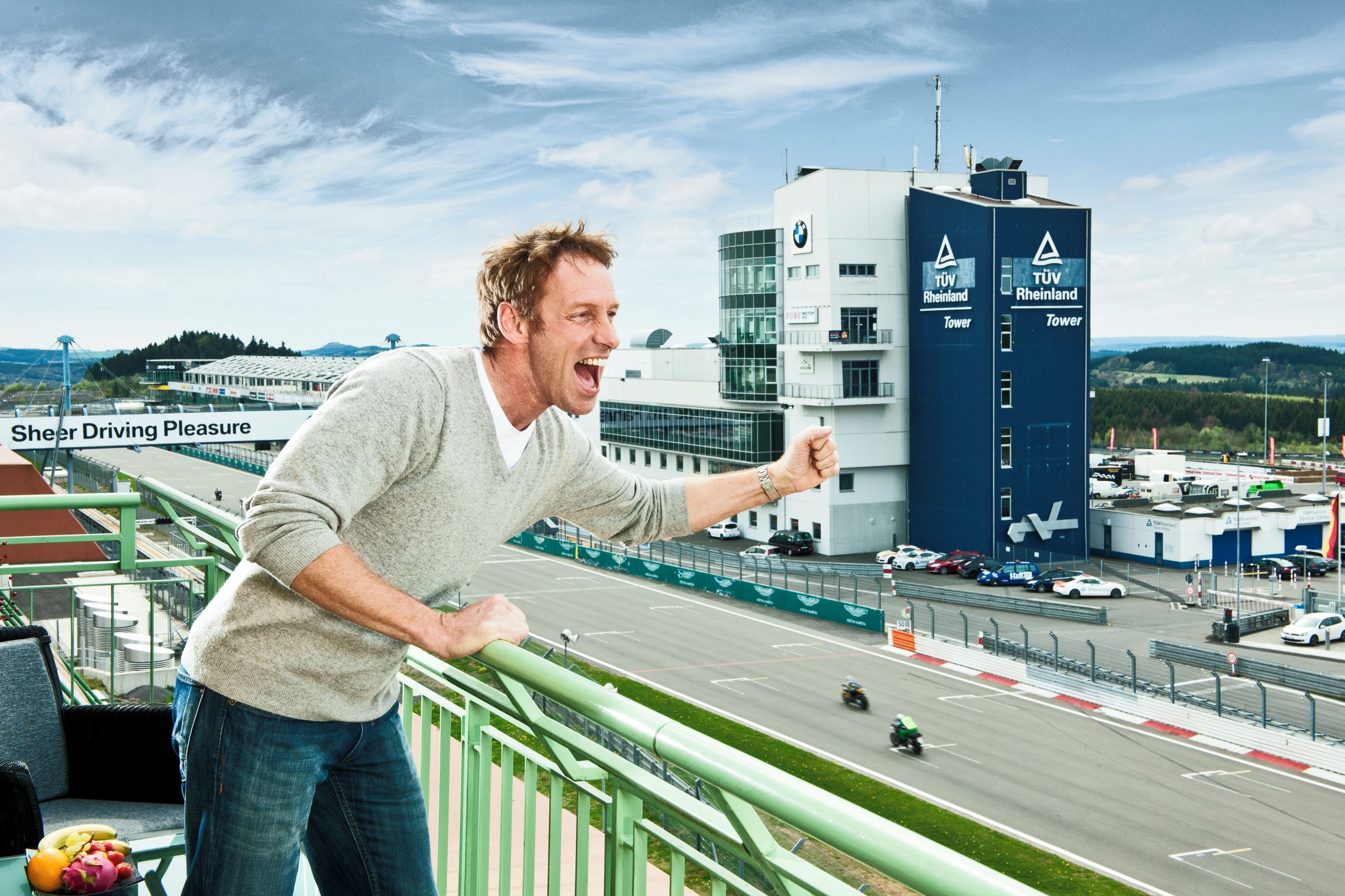 Una persona de pie en un balcón que da a una pista de carreras, con un edificio identificado como 'TÜV Rheinland' al fondo.