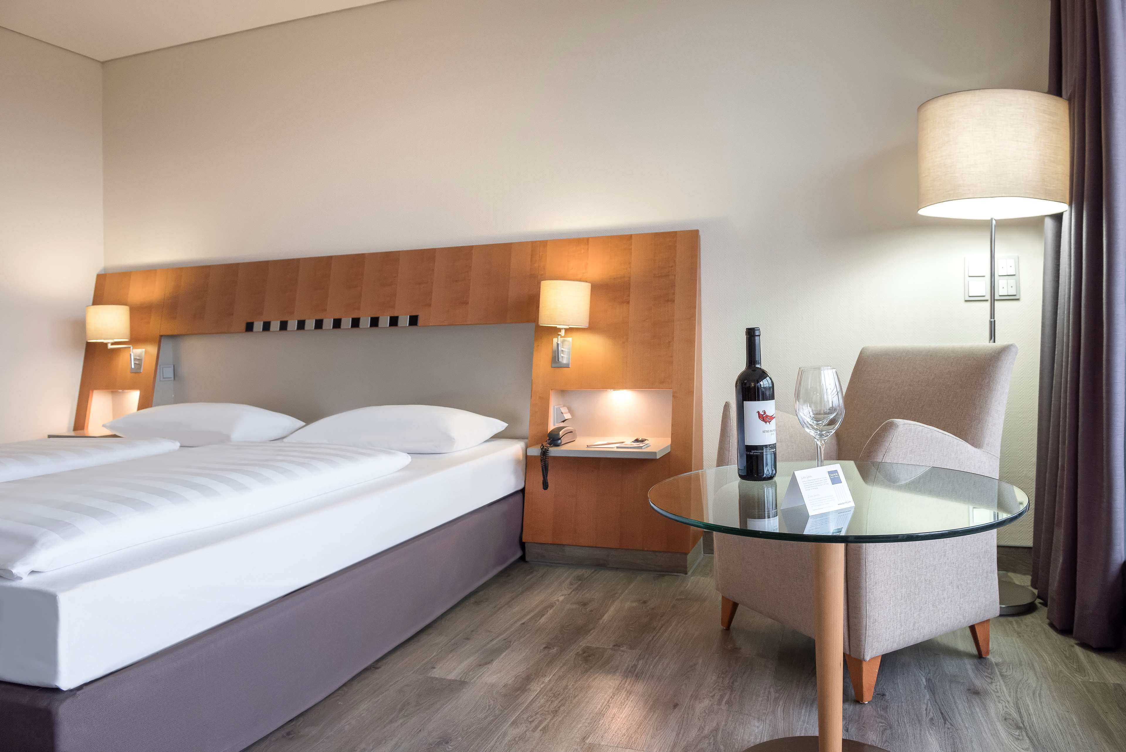 Una habitación de hotel con cama, mesitas de noche, una mesa de vidrio con una botella y vasos, y una silla.