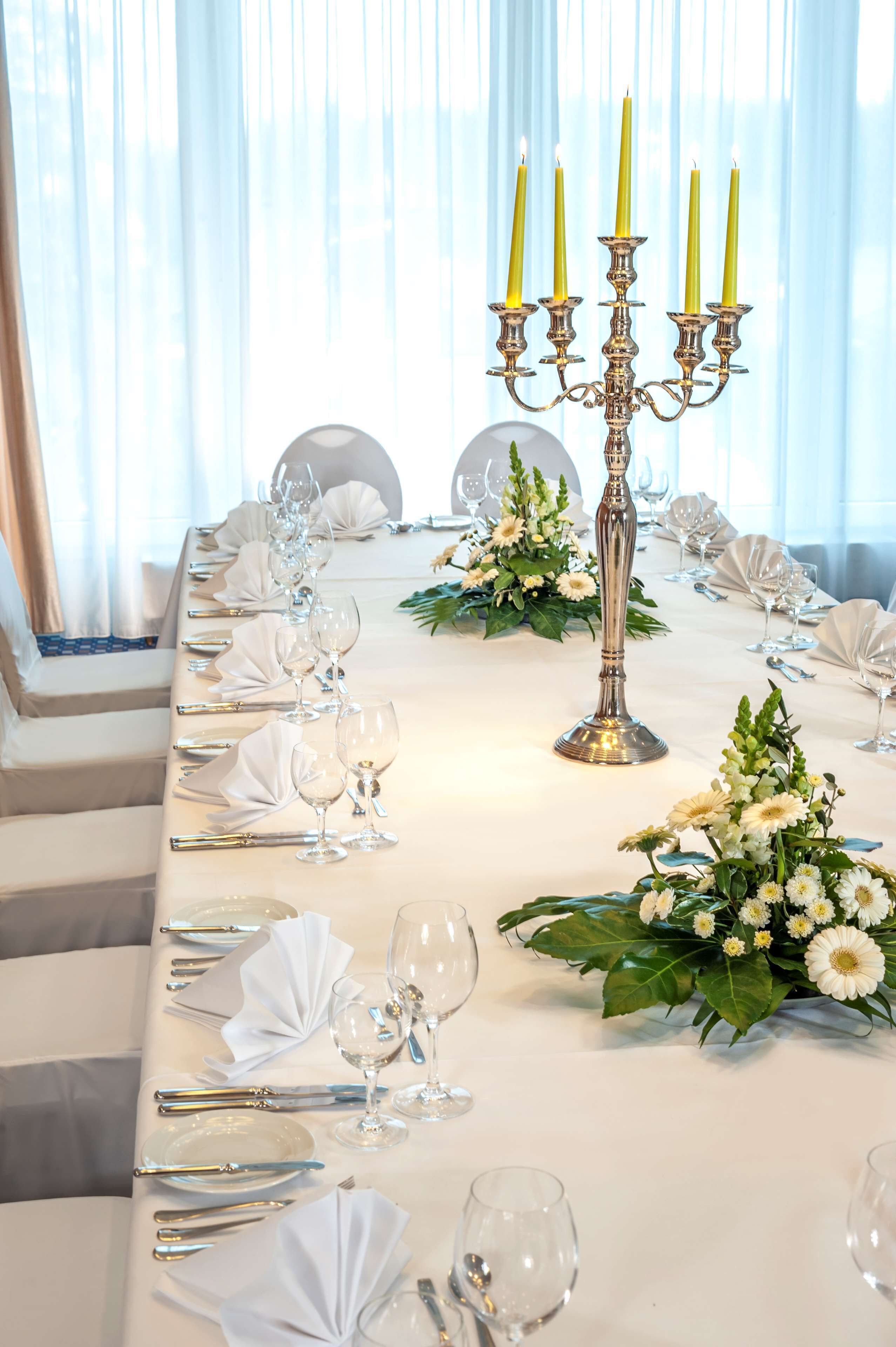 Ein langer Esstisch mit weißer Tischdecke, Besteck und Gläsern, an dessen Seiten weiße Blumenarrangements und eine hohe Kerzenleuchter mit Kerzen stehen.
