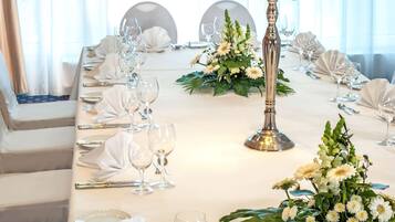 Ein langer Esstisch mit weißer Tischdecke, Besteck und Gläsern, an dessen Seiten weiße Blumenarrangements und eine hohe Kerzenleuchter mit Kerzen stehen.