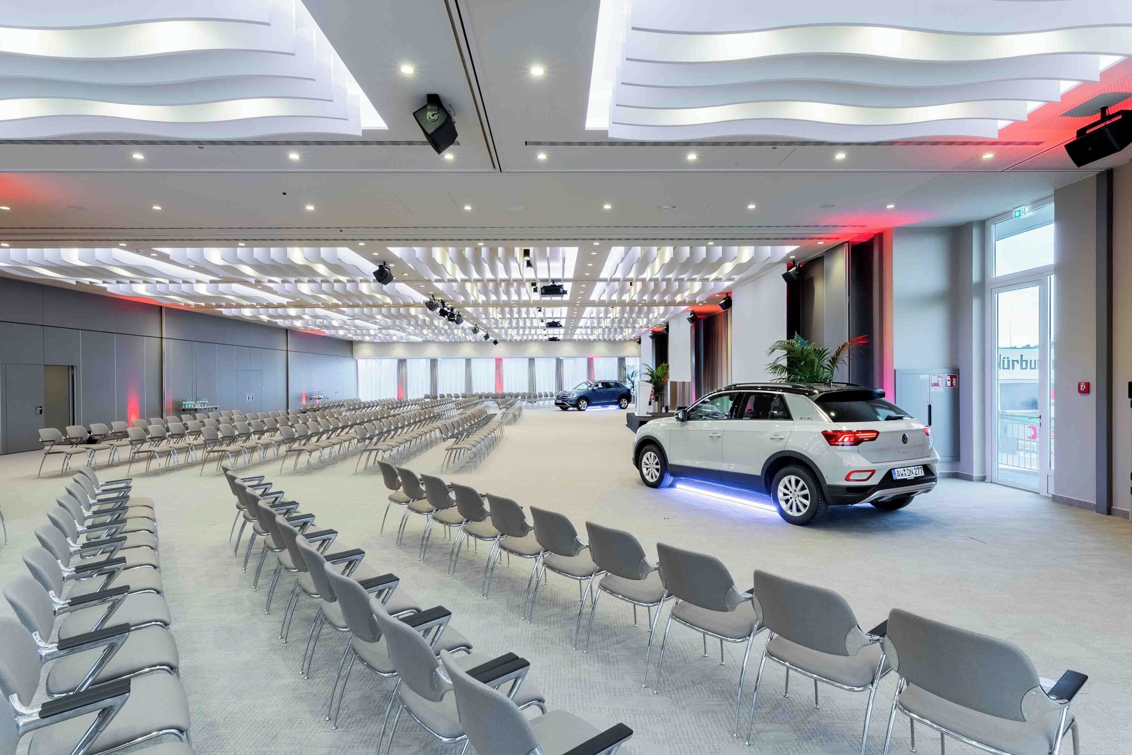 A spacious conference room with rows of chairs, a white car, and a modern ceiling design.