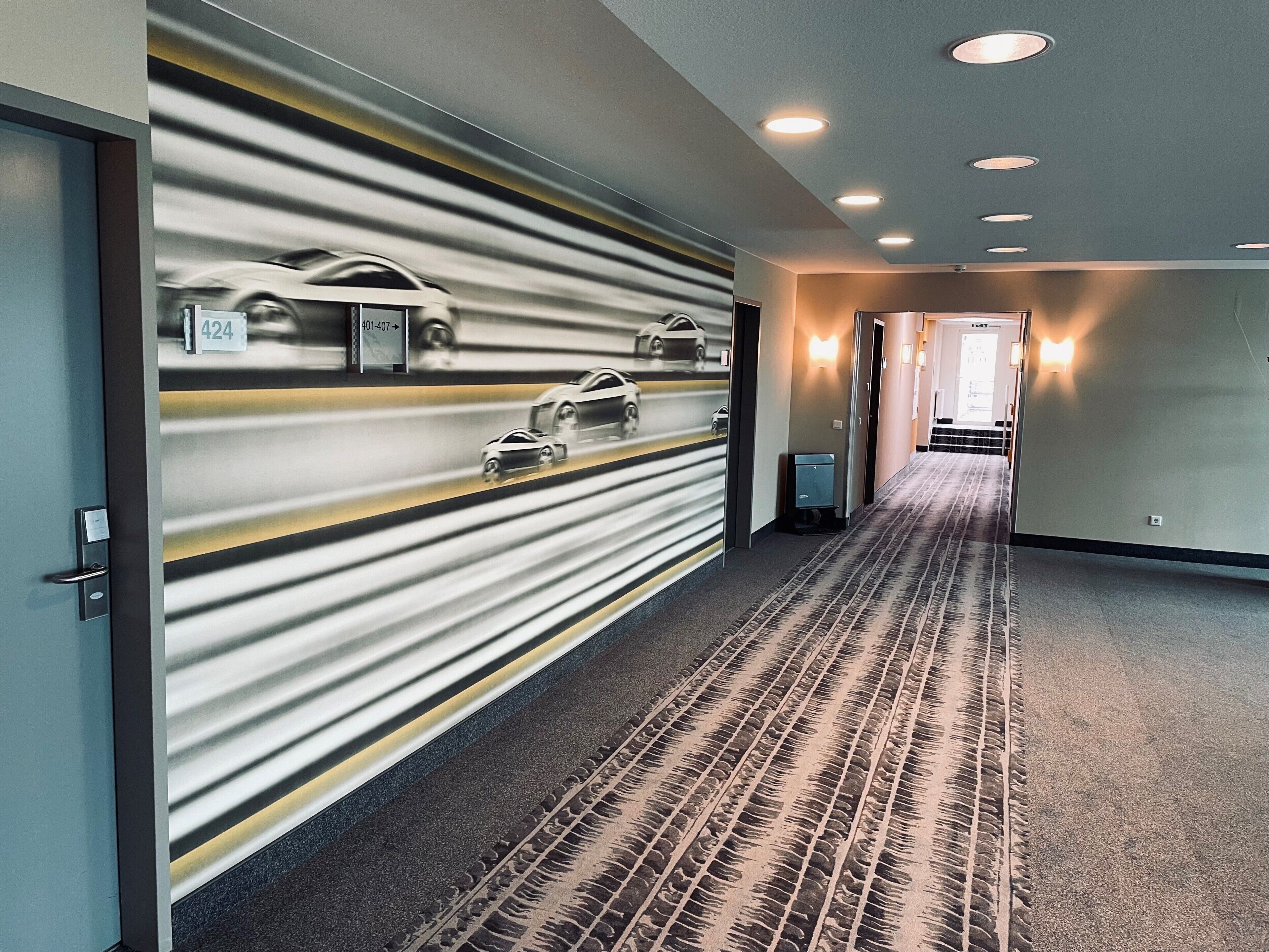 A hotel hallway with a patterned wall, a carpeted floor, and a door with a number sign.
