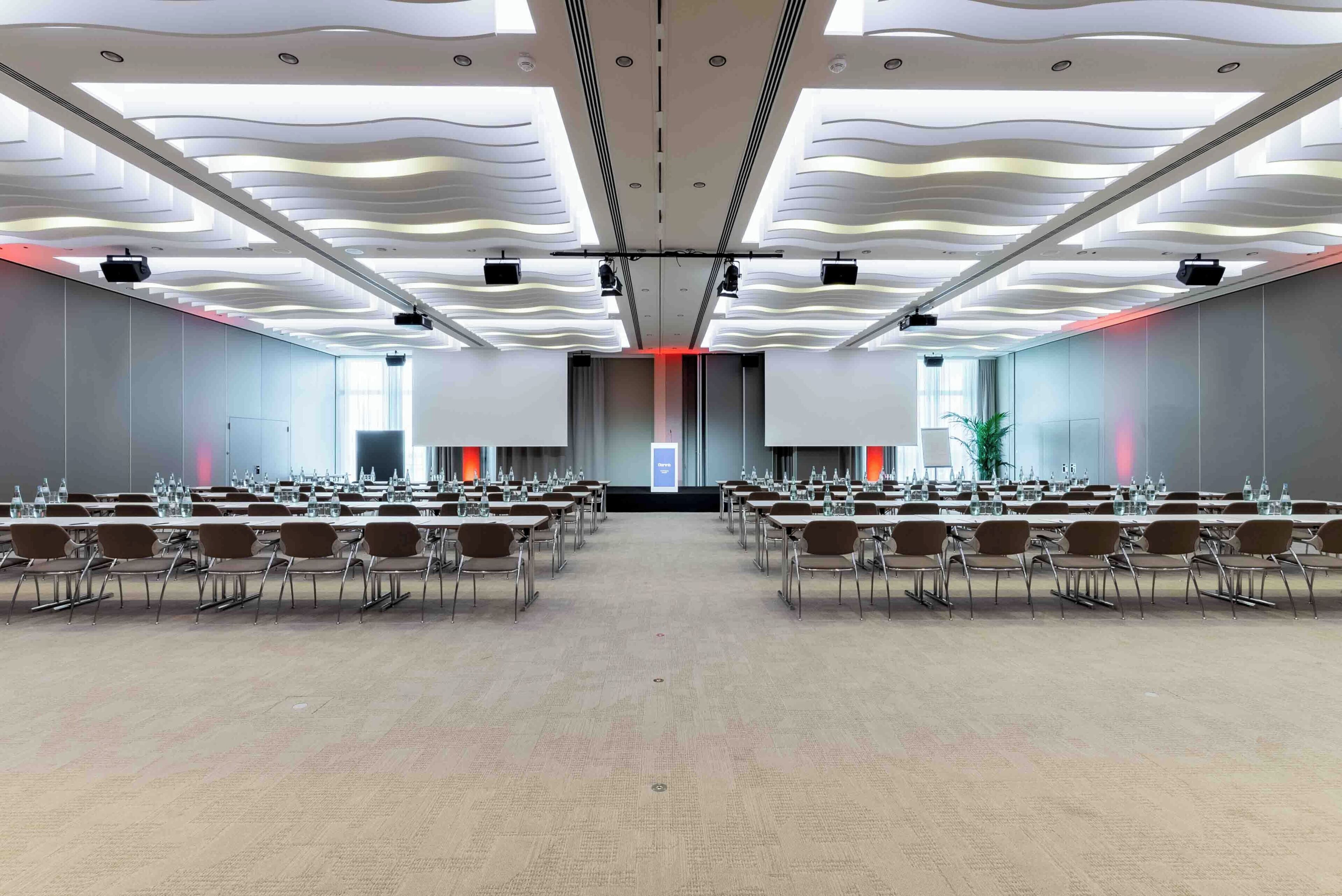 Un salón de conferencias con filas de mesas y sillas, una pantalla grande al frente y un diseño moderno de techo.