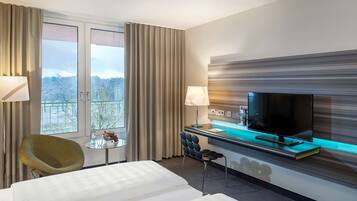Una habitación de hotel con cama, escritorio con televisor, una silla y ventana con cortinas.