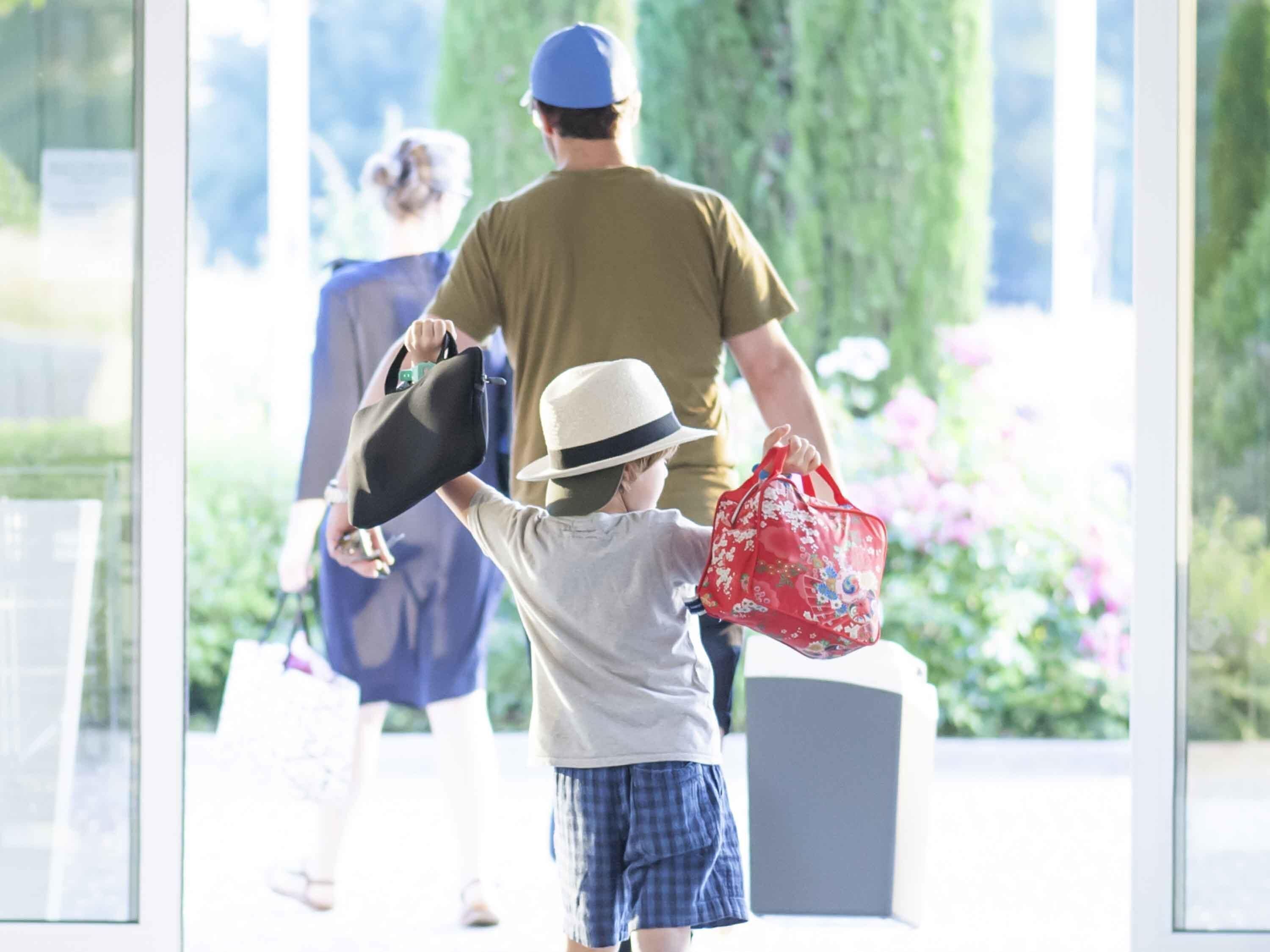 A child holding a hat and a red bag, with an adult in the background.
