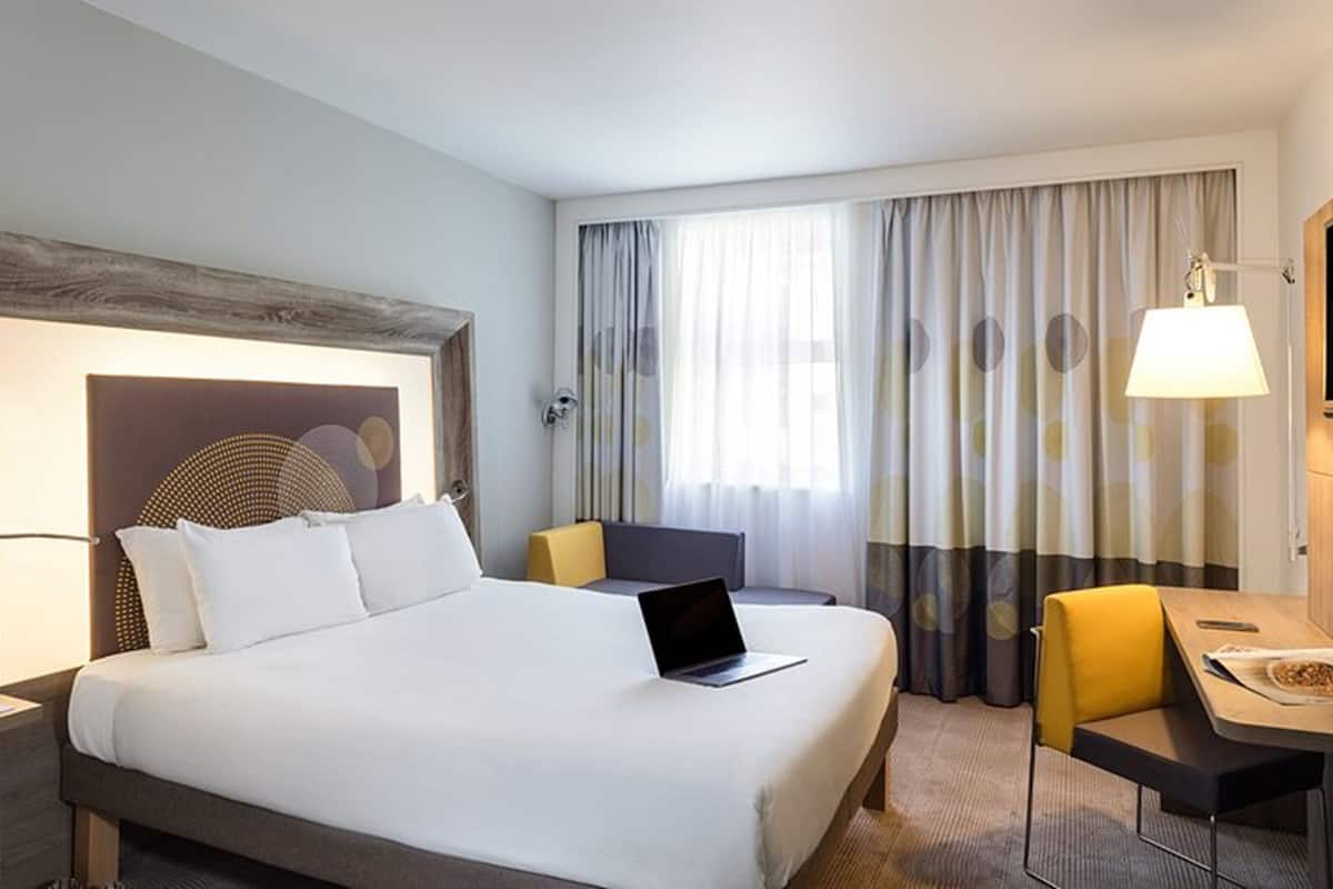 A hotel room with a large bed, a desk with a laptop, a chair, a TV, and a window with curtains.
