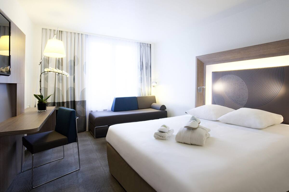 A hotel room with a bed, a desk, a chair, a lamp, and a window with curtains.
