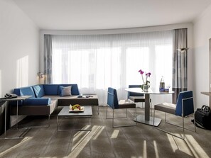 A modern hotel room with a sectional sofa, a round dining table with chairs, a small coffee table, and a large window with sheer curtains.