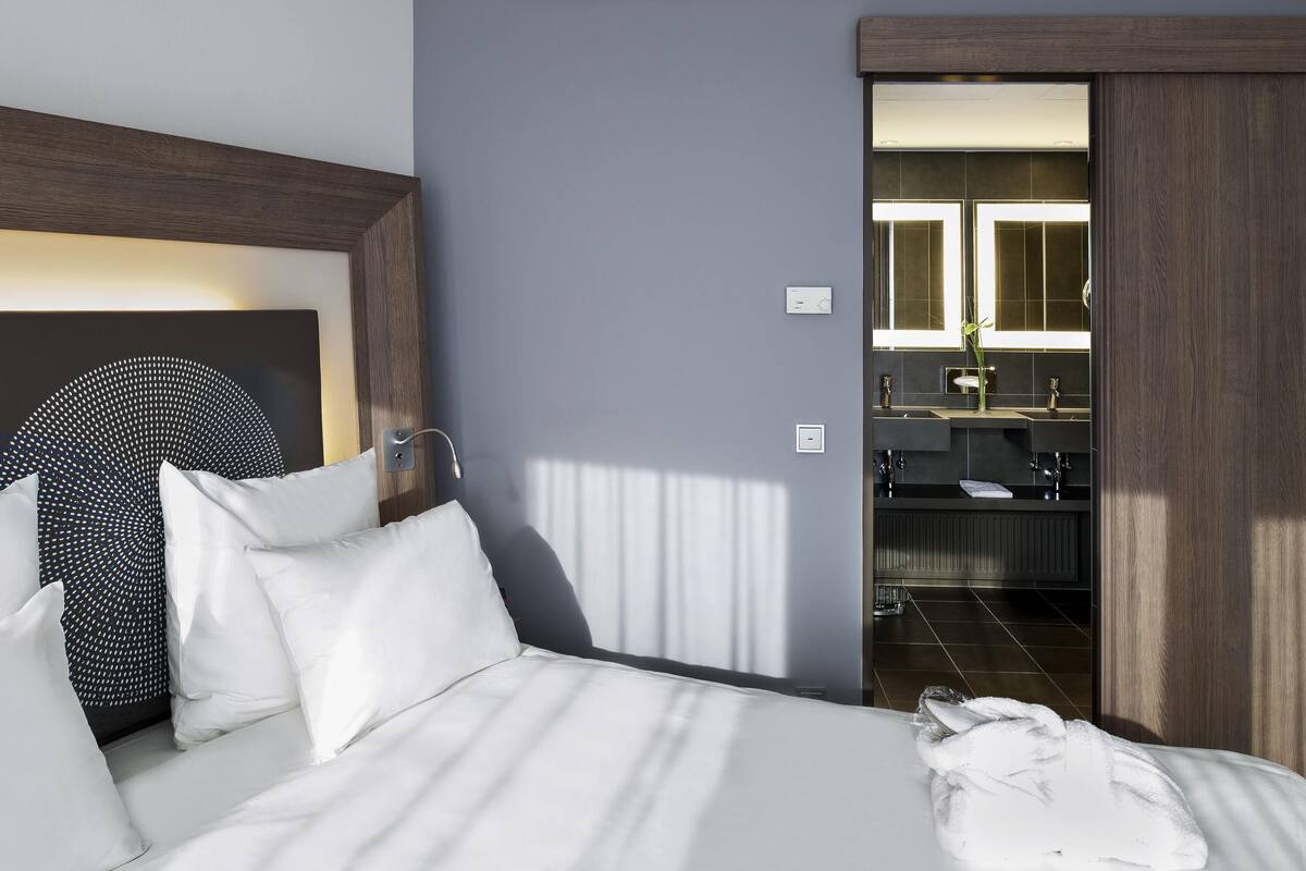 A hotel room with a bed, a bedside table, a mirror, and a bathroom visible through an open door.