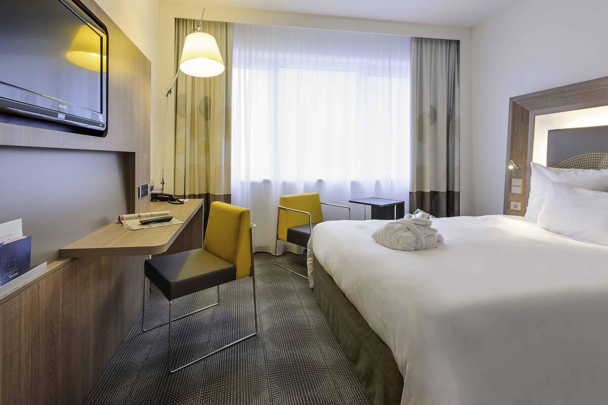 A hotel room with a bed, a desk, a chair, a TV, and a window with curtains.