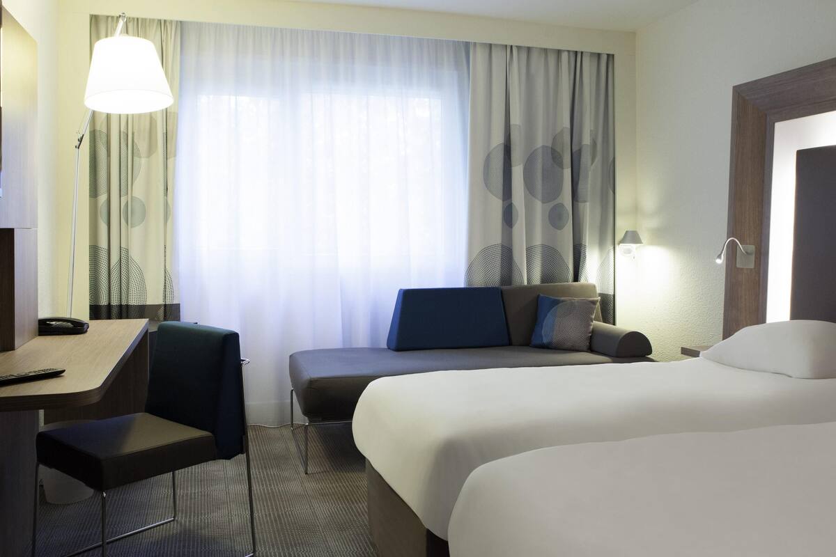 A hotel room with a bed, a desk, a chair, a lamp, and a window with curtains.