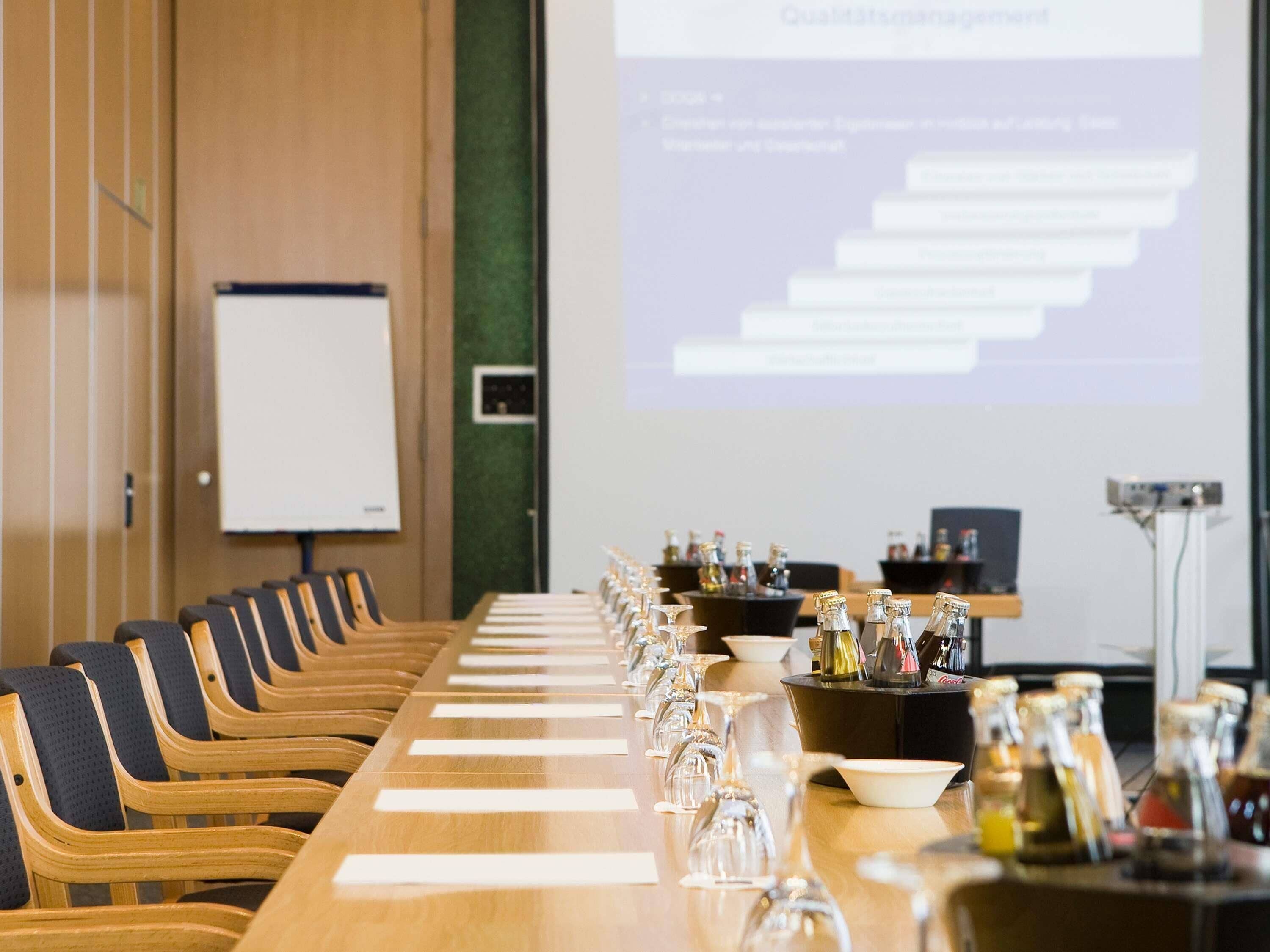 Ein Konferenzraum mit langem Tisch, gedeckten Gläsern und Flaschen, einer Whiteboard-Tafel und einer Projektionsleinwand.