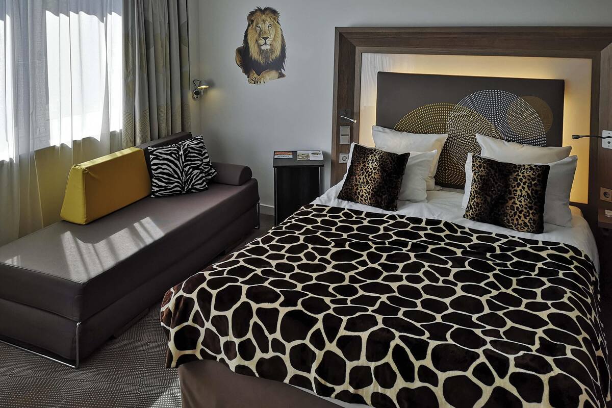 A hotel room with a large bed, a sofa, a headboard with a lion mural, and a wall-mounted lamp.