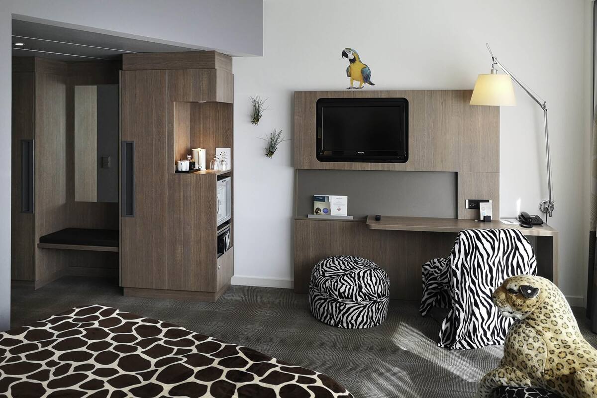 A modern hotel room with a wardrobe, a television, a desk, zebra-patterned ottoman, and a leopard-patterned sofa.