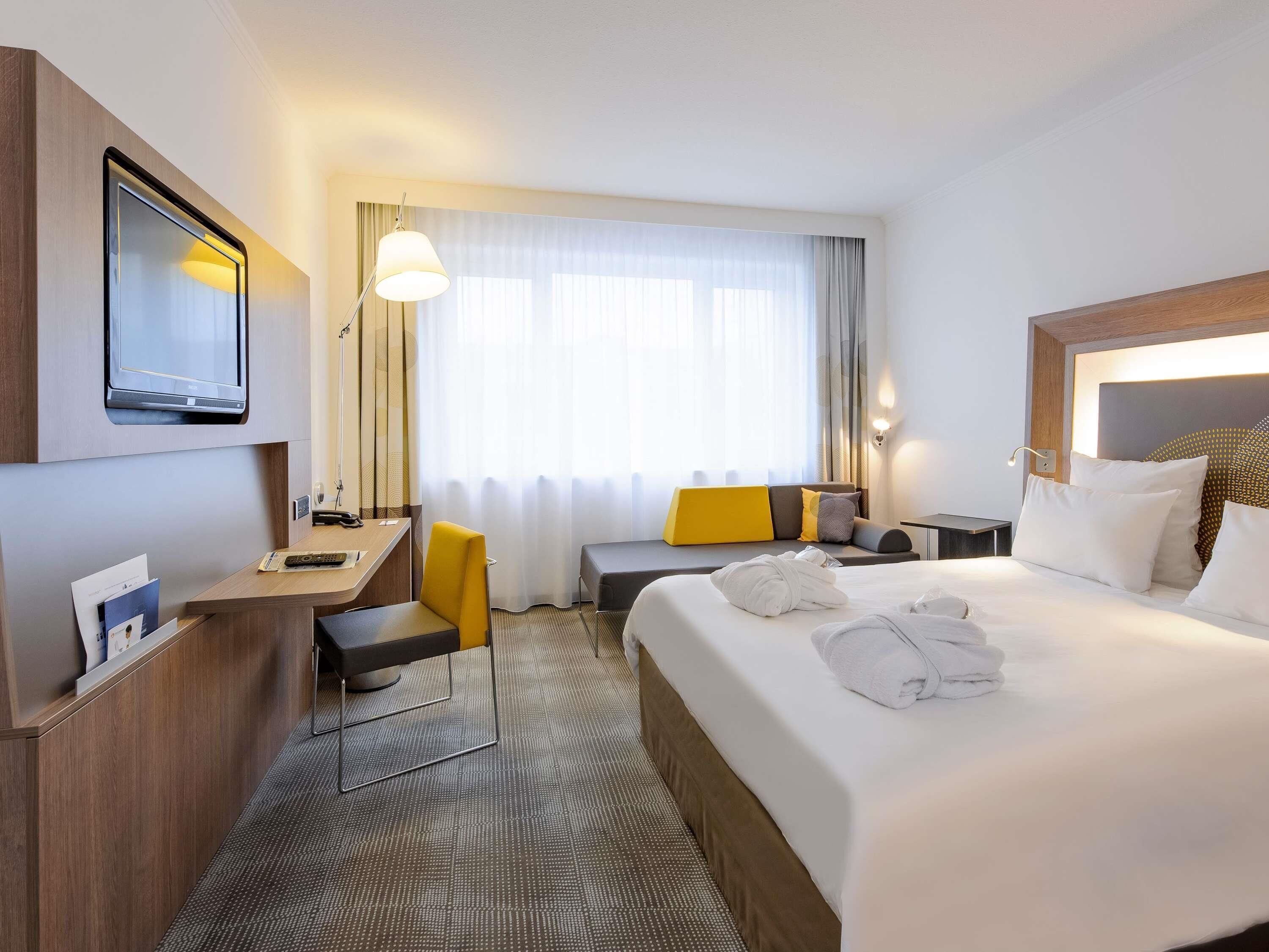 A hotel room with a bed, a desk, a chair, a TV, a lamp, and a window with curtains.