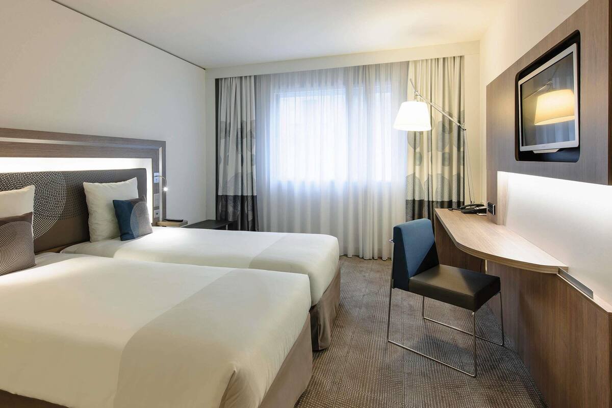 A hotel room with two beds, a desk, a chair, a lamp, and a TV.