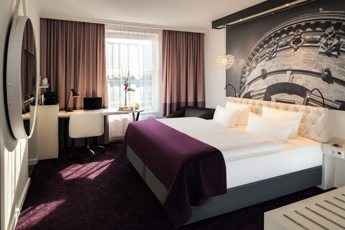 Een hotelkamer met een groot bed, een bureau met een computer, een stoel en een raam met gordijnen.