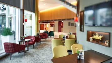 A hotel lobby with a carpeted floor, red and yellow chairs, a wooden table with a vase, and a TV on the wall.