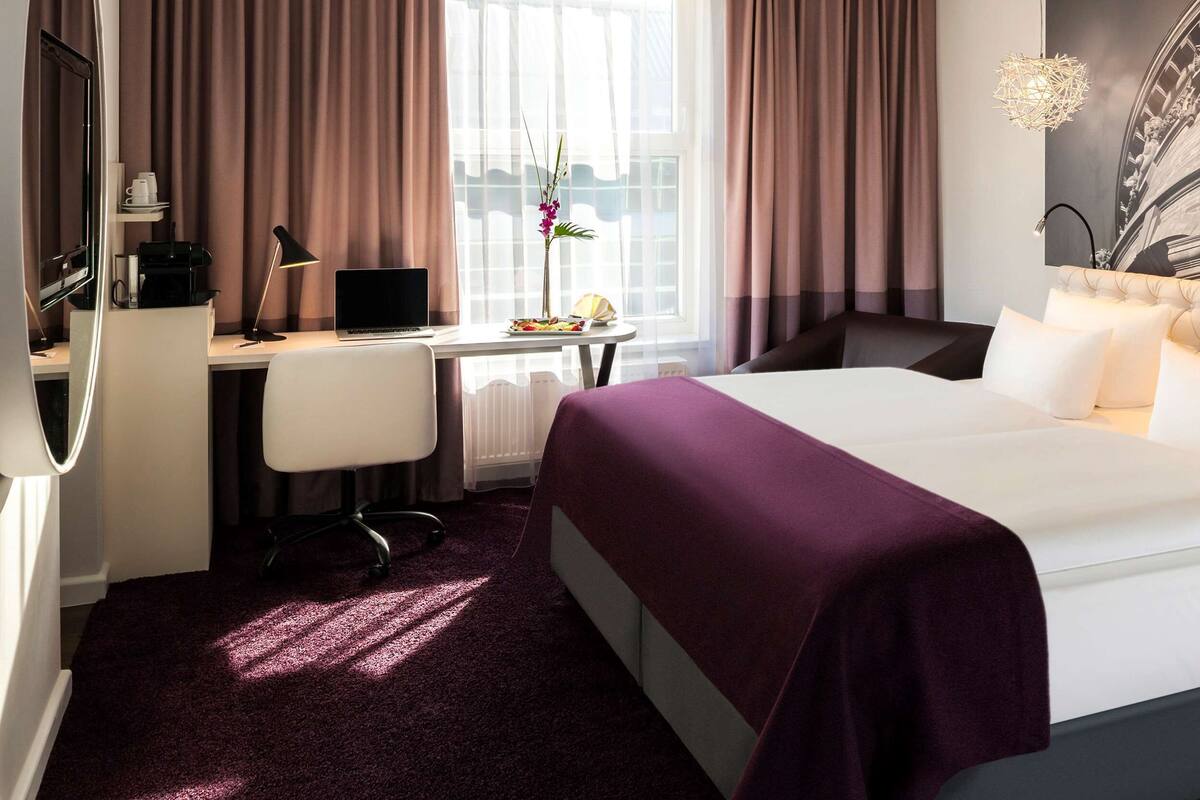 Una habitación de hotel con cama, escritorio, silla y ventana con cortinas.