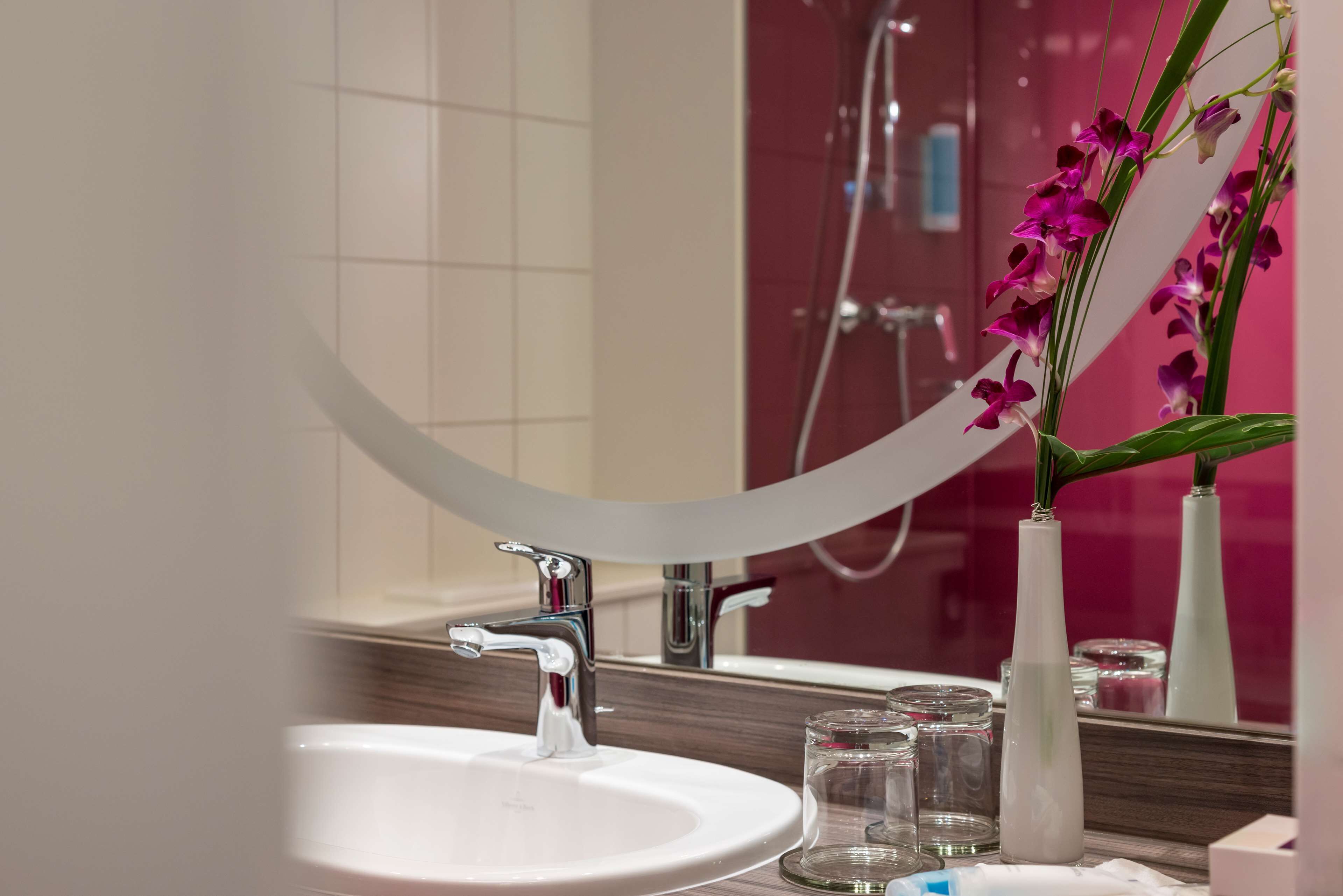 Une salle de bain avec un lavabo blanc, un miroir et un vase contenant des fleurs violettes.