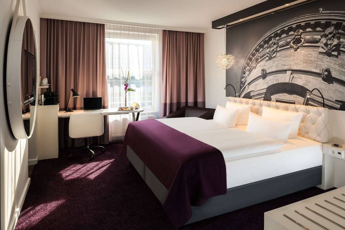 Una habitación de hotel con una cama grande, un escritorio con computadora, una silla y una ventana con cortinas.