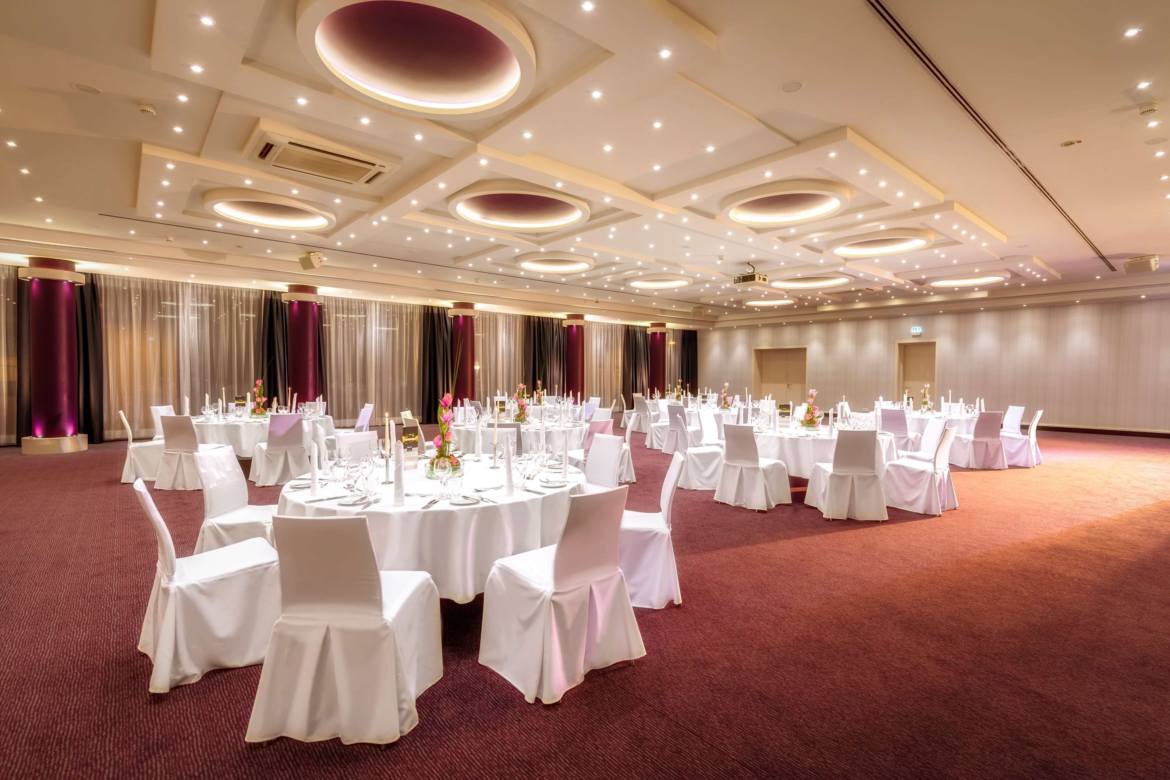 Un salón de banquetes con mesas redondas, manteles blancos y sillas, que incluye una alfombra roja y un diseño moderno del techo.
