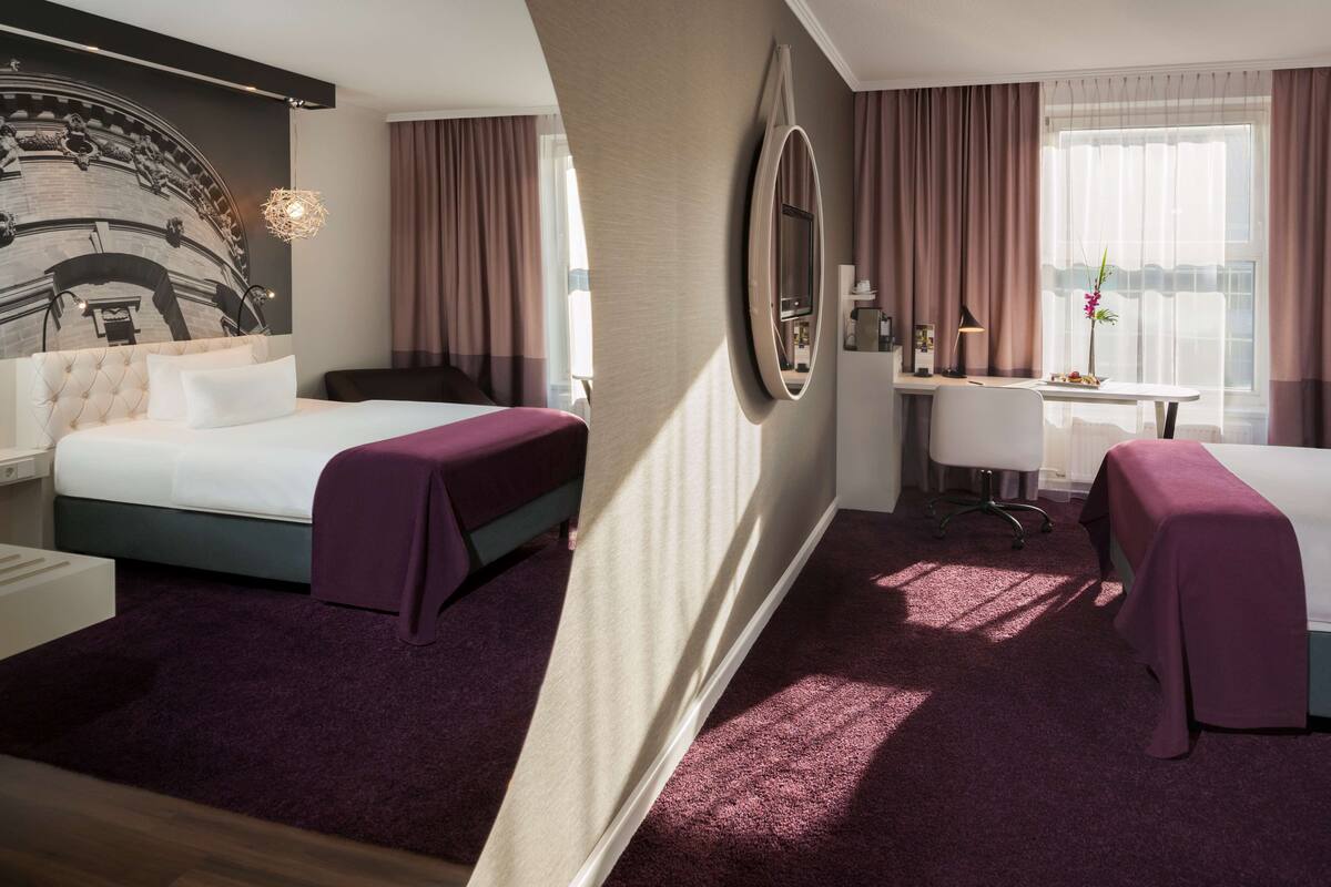 Habitación de hotel con cama, escritorio, silla, televisor y ventana grande con cortinas.