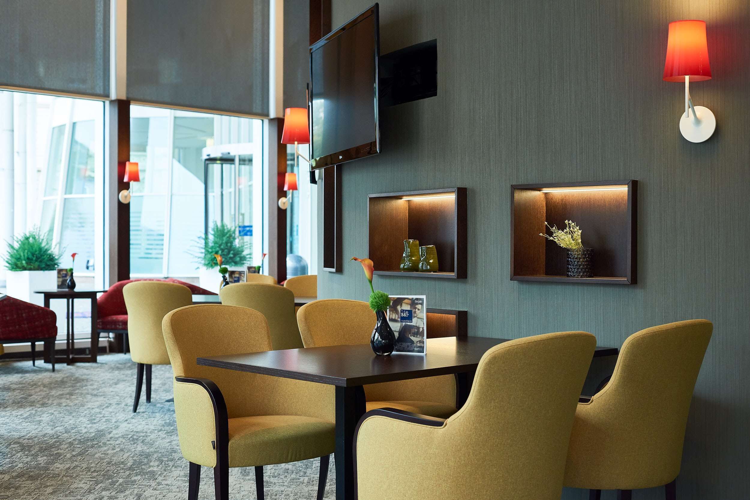 Un vestíbulo de hotel con diseño moderno, que incluye un área de comedor con sillas amarillas, una mesa con un florero y televisores instalados en la pared.