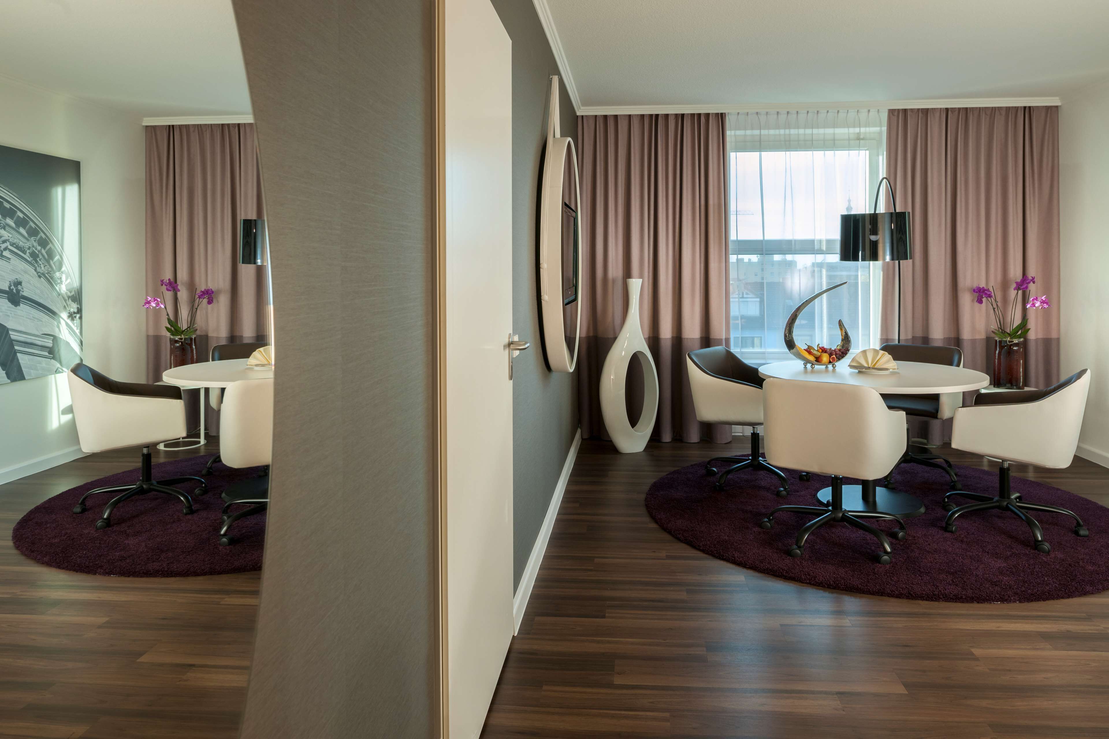 A modern hotel room with a dining area, wooden flooring, and a large window with curtains.