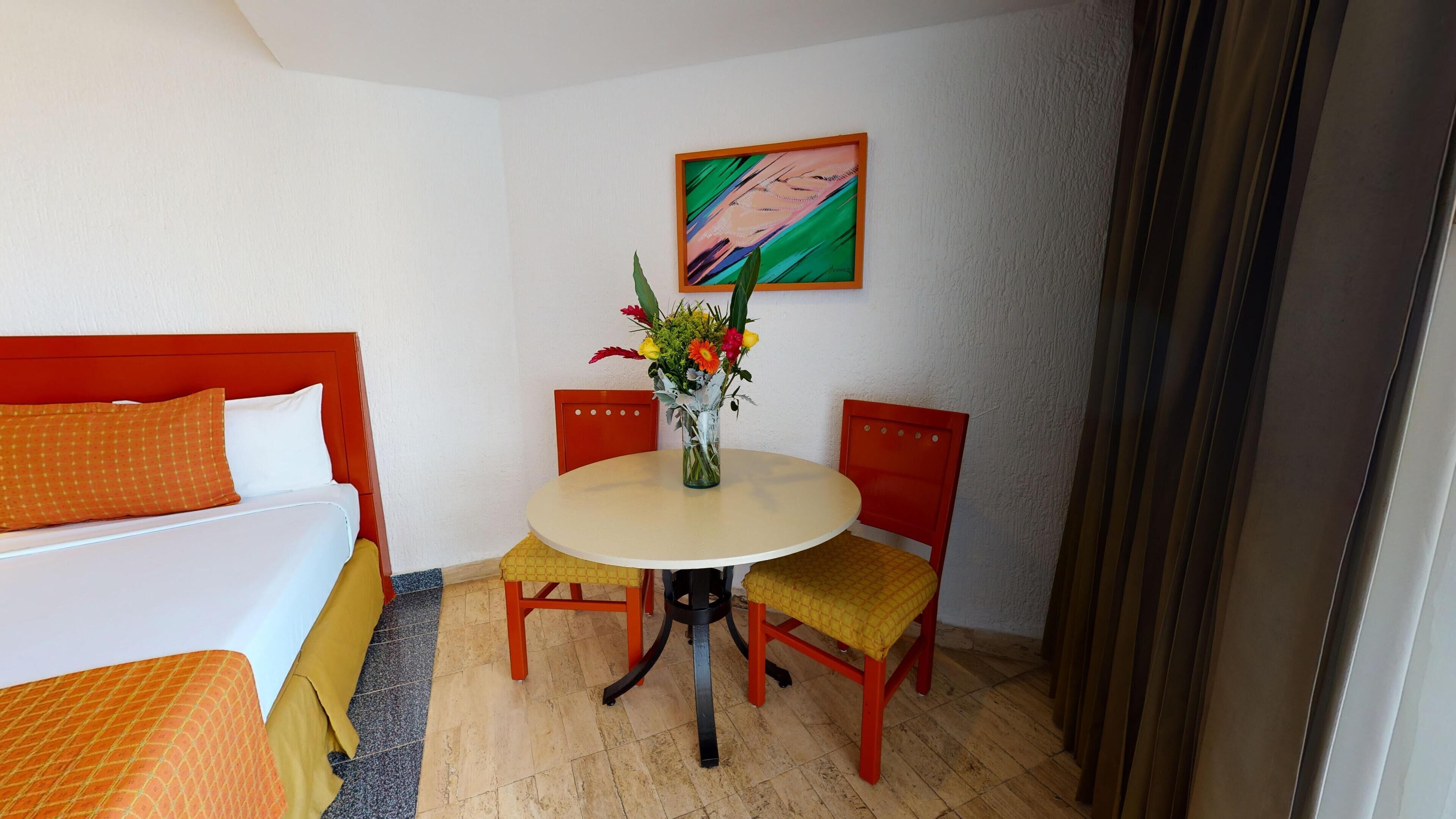 Habitación de hotel con cama, una mesita redonda con un florero y dos sillas, además de un cuadro en la pared.