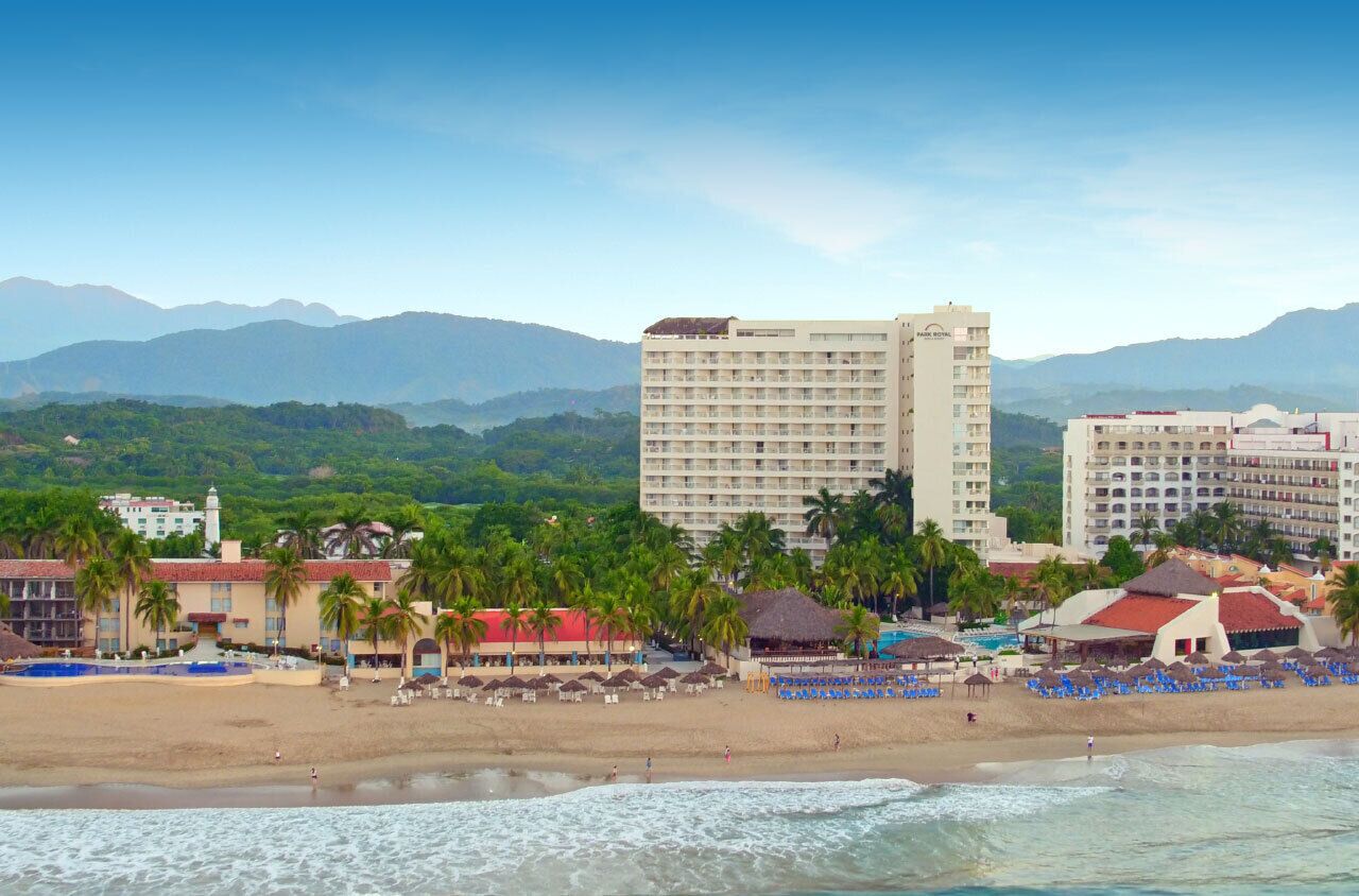 Un hotel frente a la playa con varias construcciones, una playa de arena y palmeras.