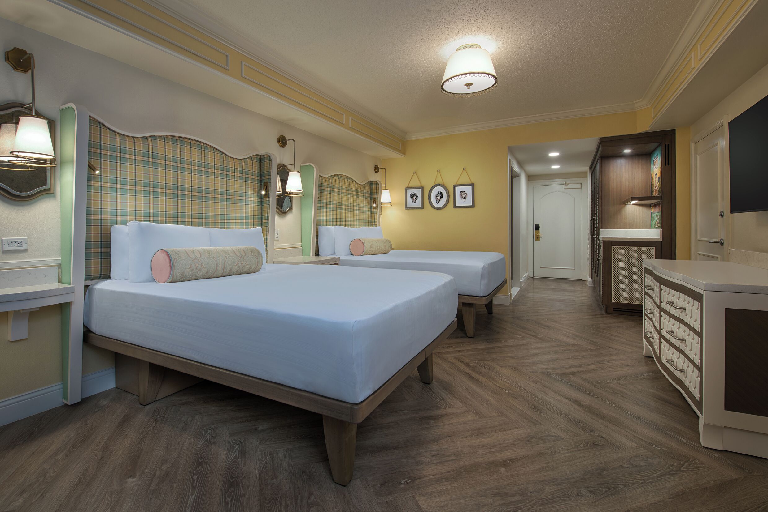 A hotel room with two beds, a vanity with a mirror, wall-mounted lamps, and a herringbone-patterned floor.