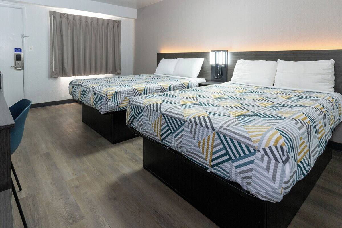 Hotelkamer met twee bedden, elk voorzien van een beddekleed met dessin en witte kussens. Een bureau met stoel, een kleine prullenbak en een deur met slot zijn ook aanwezig.