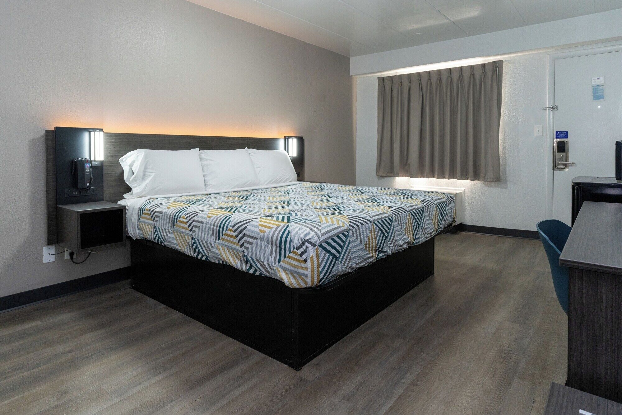Habitación de hotel con cama, mesita de noche, lámpara y ventana con cortinas.