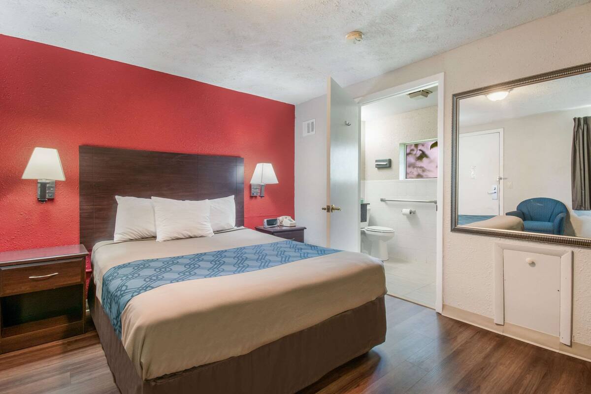 Habitación de hotel con una cama grande, dos lámparas de noche, una pared de acento roja, piso de madera, baño con inodoro y lavamanos, y un espejo con marco decorativo.