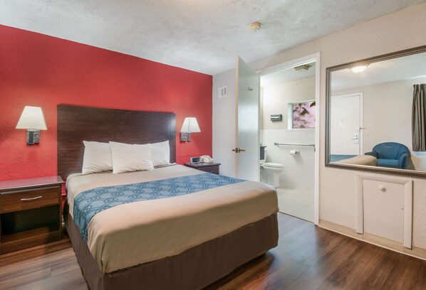 Habitación de hotel con una cama grande, dos lámparas de noche, una pared de acento roja, piso de madera, baño con inodoro y lavamanos, y un espejo con marco decorativo.
