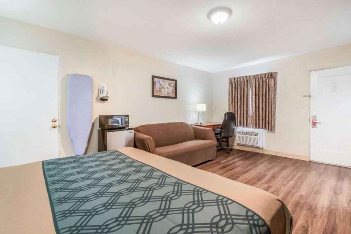 Habitación de hotel con una cama, un escritorio con computadora, una silla, una lámpara y una ventana con cortinas.