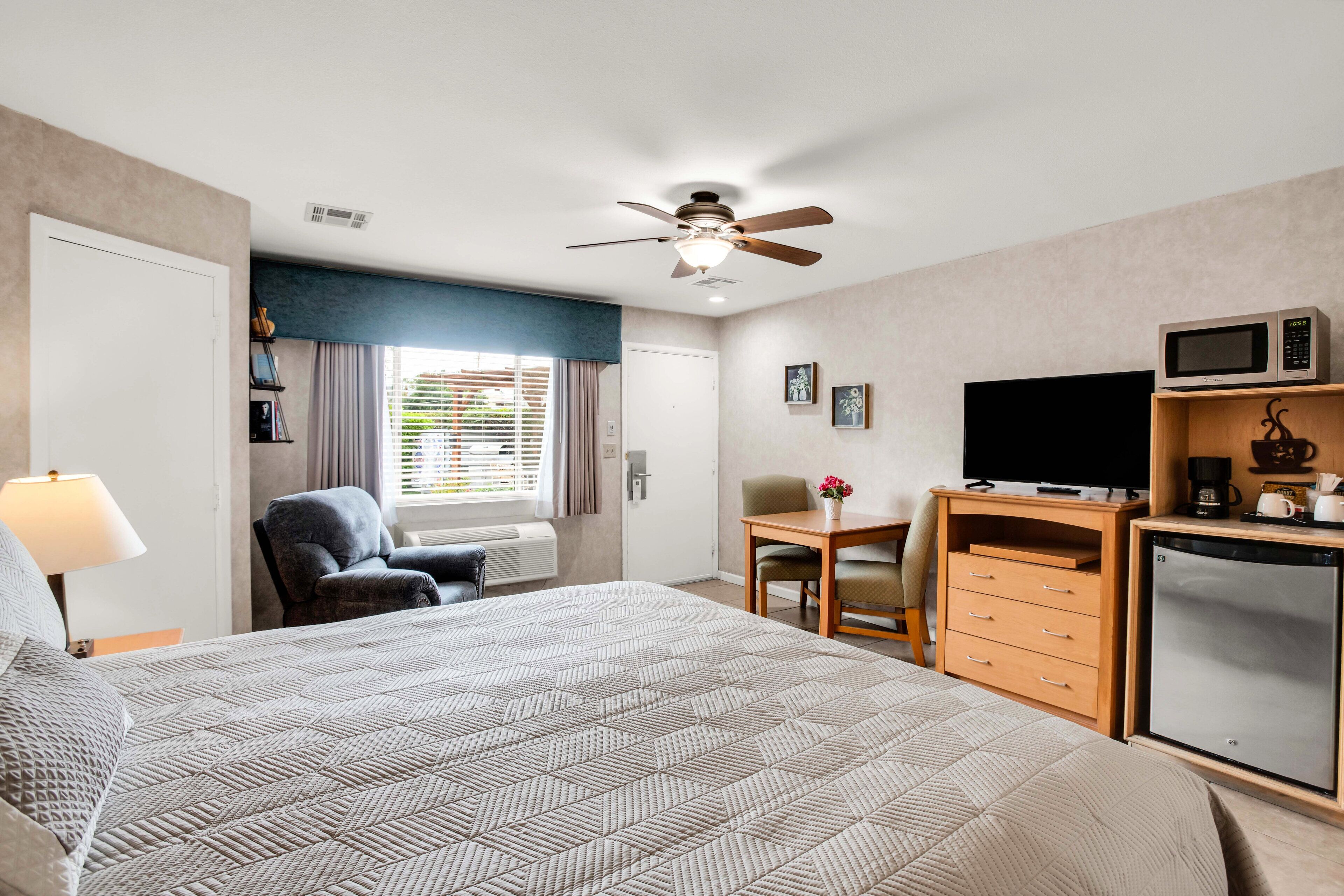 A hotel room with a bed, desk, chair, microwave, refrigerator, and a ceiling fan.