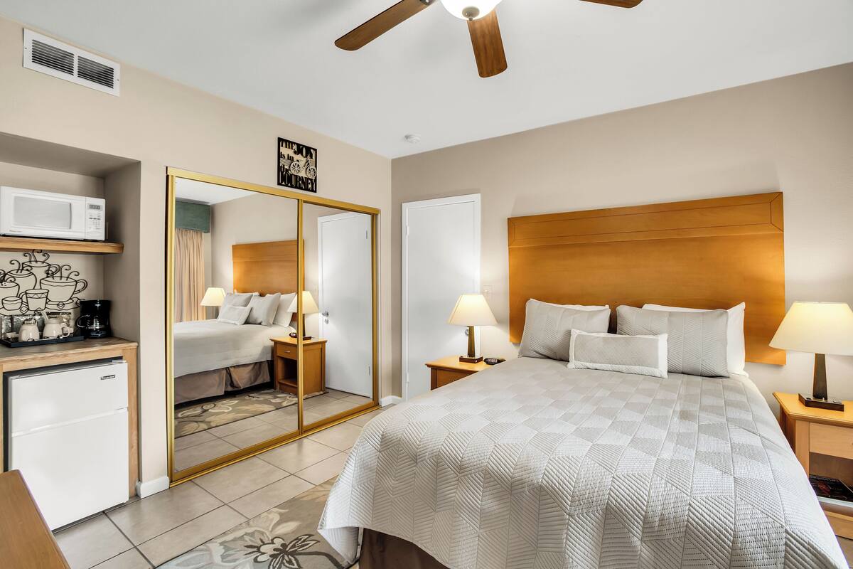 A hotel room with a large bed, a wooden headboard, a ceiling fan, a microwave, a small refrigerator, and a mirror with a door.