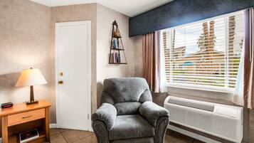 Une pièce avec un fauteuil gris, une table de chevet en bois avec une lampe, une bibliothèque, une porte blanche et une fenêtre avec des stores.