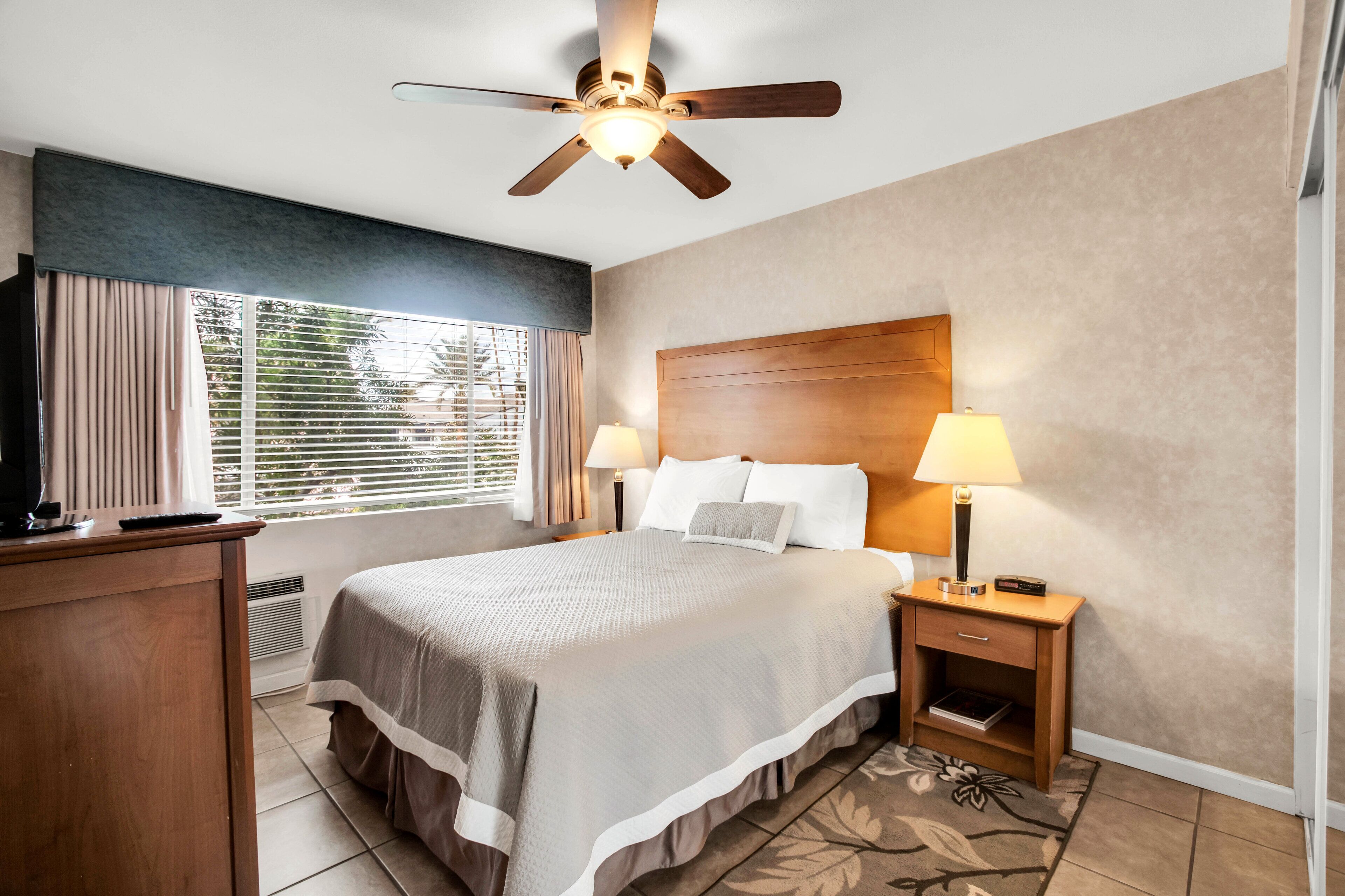 A hotel room with a bed, two bedside lamps, a ceiling fan, a window with blinds, a desk, and a nightstand.