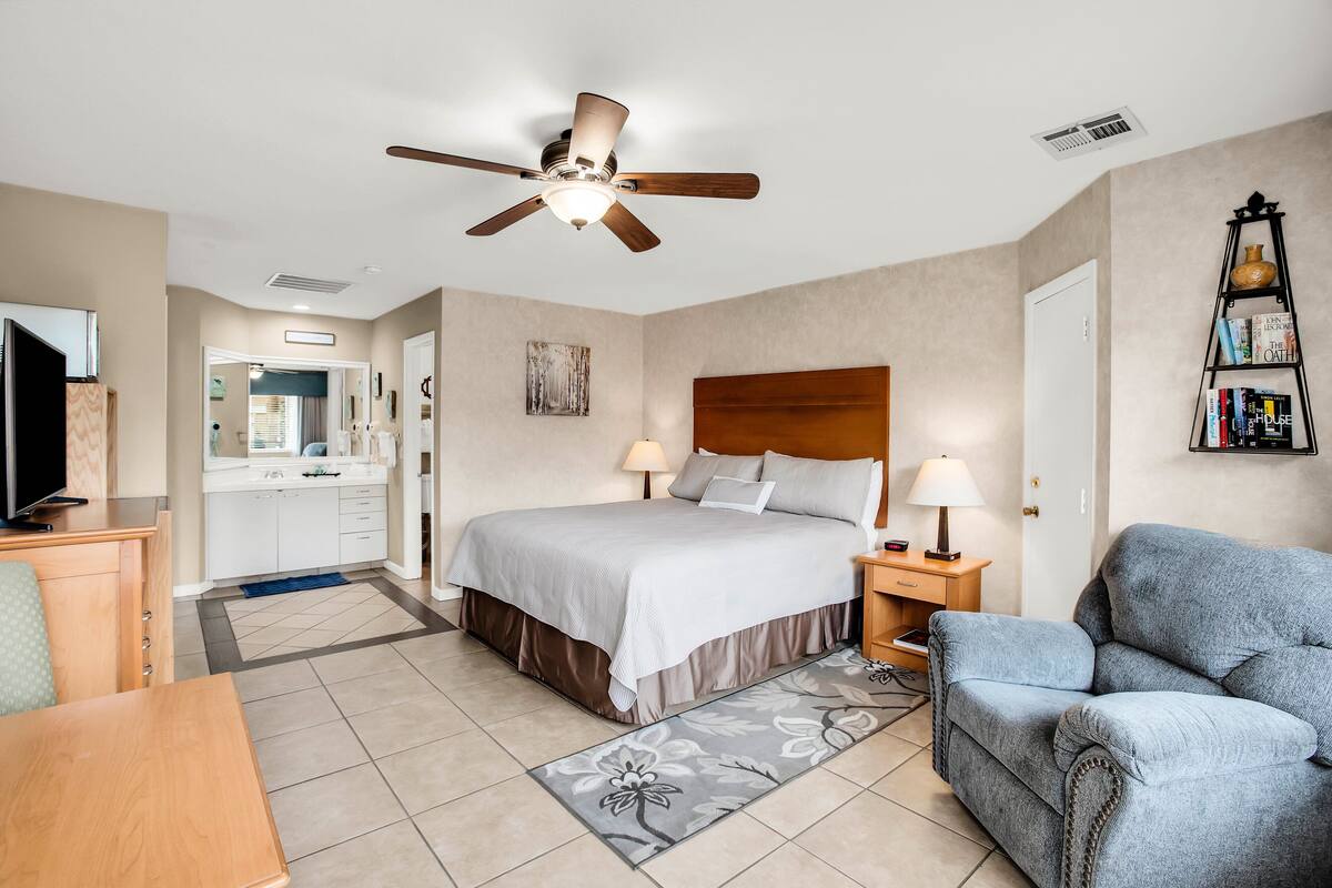 A bedroom with a bed, bedside lamps, a ceiling fan, a bookshelf, and a view into the bathroom.